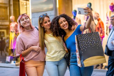 Üç farklı etnik çeşitlilikte genç kadın alışveriş merkezinde selfie çekiyorlar.