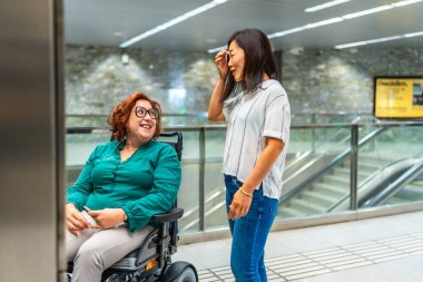 Gülümseyen Kafkas yetişkin engelli kadın ve Asyalı bayan arkadaş metro asansörünü bekliyorlar.