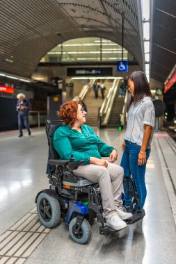 Tekerlekli sandalyedeki engelli bir kadının metro istasyonunda bekleyen Asyalı bir bayan arkadaşla konuşurken çekilmiş dikey fotoğrafı.