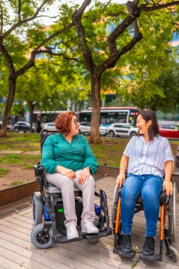 Tekerlekli sandalyedeki iki etnik engelli kadın şehir parkında konuşuyor.