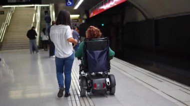 Kafkasyalı, tekerlekli sandalyedeki yetişkin bir kadının ve metro istasyonunda yürüyen Asyalı bir arkadaşın arka görüntüsü.