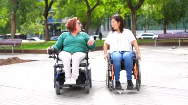 Şehir parkında tekerlekli sandalyeyle konuşan engelli çok ırklı, mutlu bayan arkadaşlar.