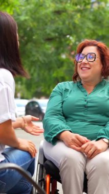Engelli mutlu kadın, şehir bankında oturan Asyalı bir kadınla konuşuyor.