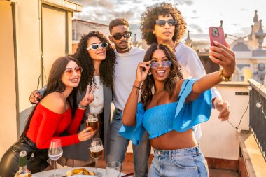 Kadın, çok ırklı arkadaşlarıyla bir çatı partisinde gün batımında şarap ve atıştırmalıklarla selfie çekiyor.