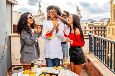 Bir adamın gözlerini kapayan ve çatıda doğum günü şarkısı söyleyen çok ırklı arkadaşlar.