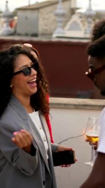 Çok ırklı insanlar çatıda akşam partisini kutluyor şarap içiyor ve sohbet ediyorlar.