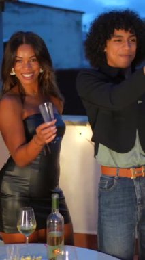 Çok ırklı arkadaşlarla çatıda şampanya şişesi açıp kutlama yapan mutlu bir adam.