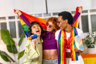 Çok ırklı ve cinsiyetli arkadaşlar evde karaoke söylüyor ve Igbt gökkuşağı bayrağı dikiyorlar.