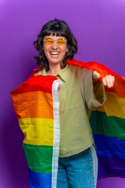 lgbt gökkuşağı bayrağına sarılı, ikili olmayan mutlu bir insanın mor arkaplanlı stüdyo portresi