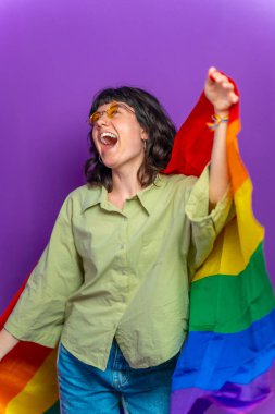 Stüdyo portresi mor arka planda kot elbiseli ve güneş gözlüklü lgbt raibow bayrağıyla dans eden genç bir kadın.