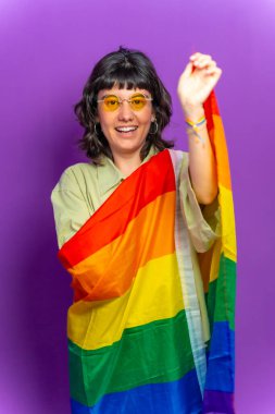 Lgbt gökkuşağı bayrağıyla kaplı mutlu bir lezbiyen kadının mor arkaplanlı stüdyo portresi kameraya gülümsüyor.