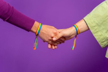 Mor arkaplanlı bir stüdyo fotoğrafı ve lgbt gökkuşağı bileziği takan iki lezbiyen kadının yakın çekimi.