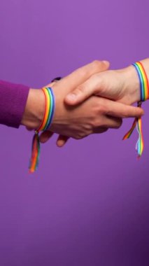 LGBT gökkuşağı bileziği takan tanınmayan lezbiyen bir çiftin ellerinin mor arkaplanlı dikey fotoğrafı.