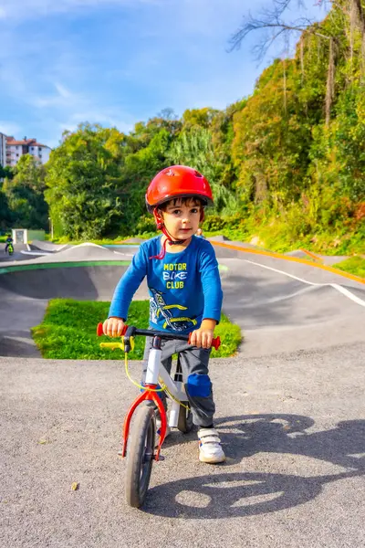 Kask takan genç bir çocuk sokakta bisiklet sürüyor. Çocuk mavi bir gömlek ve gri pantolon giyiyor.