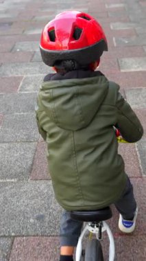 Genç bir çocuk kaldırımda bisiklet sürüyor. O bir kask ve yeşil ceket giyiyor.