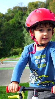 Kask takan genç bir çocuk sokakta bisiklet sürüyor. Çocuk mavi bir gömlek ve gri pantolon giyiyor.