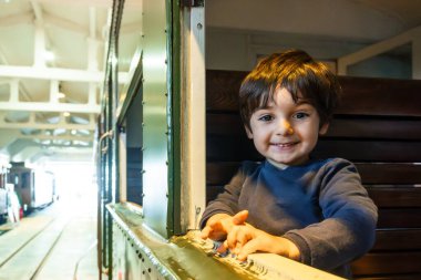 Genç bir çocuk gülümsüyor ve bir trenin penceresinden dışarı bakıyor. Tren istasyonda ve çocuk bankta oturuyor.