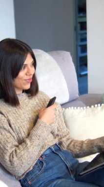 Bir kadın kanepede oturuyor ve televizyon izliyor. Elinde bir uzaktan kumanda tutuyor.