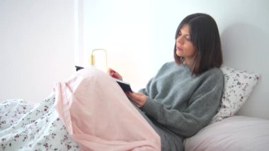 Bir kadın yatakta yatarken bir kitap okuyor. Gri bir kazak giyiyor ve üzerinde pembe bir battaniye var. Rahatlama ve rahatlama kavramı