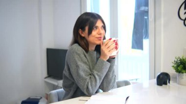 Bir kadın önünde bir kupa kahveyle masada oturuyor. Gri bir kazak giyiyor ve kırmızı tırnakları var. Sahne rahat ve rahatlatıcı. Kahvesini içen kadınla.