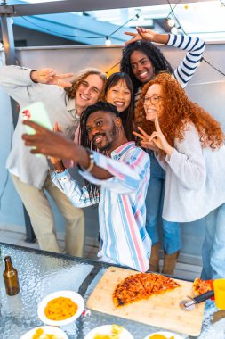 Günbatımında çatıdaki bir partide, gülümseyen Afrikalı bir adamın çok ahlaklı arkadaşlarıyla selfie çekerken çekilmiş dikey fotoğrafı.
