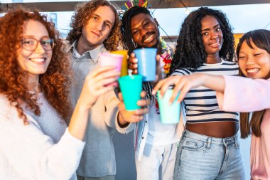 Gün batımında bir teras partisinde kameraya kadeh kaldıran çok ırklı genç arkadaşlar.