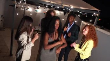 Gece partisinde terasta arkadaşlarıyla dans eden şık bir gece elbisesi ve güneş gözlüğü takan Afrikalı kadın.