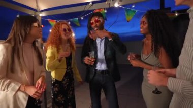 Çok ırklı, şık giyinmiş genç insanlar terasta gece partisinde sohbet ediyorlar.