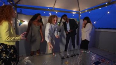 Çok ırklı, zarif bir arkadaş grubu çatıdaki bir partide şampanyayla kameraya kadeh kaldırıyor.