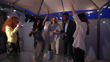 Çok ırklı, zarif bir arkadaş grubu çatıdaki bir partide şampanyayla kameraya kadeh kaldırıyor.