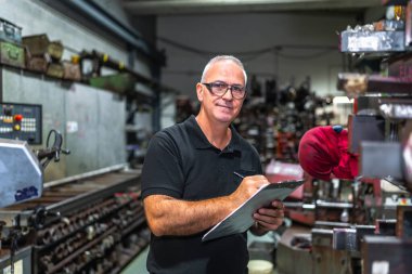 Olgun bir erkek işçi, kameraya ciddi ciddi bakarken elinde pano olan metal bir fabrikada envanter sayımı yapıyor.