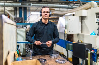 Metal parçalarının boyutlarını ölçmek için mikrometre kullanan bir adam metal fabrikasında koruyucu kulaklık takıyor.