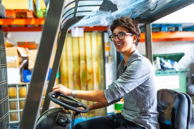 Metalurjik bir fabrikada forklift kullanan kısa saçlı ve gözlüklü beyaz bir kadının yan görüntüsü.