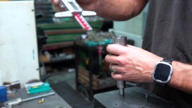 Beyaz erkek operatör mikrometre kullanarak fabrikadaki metal parçaları üzerinde kalite kontrolü yapıyor.