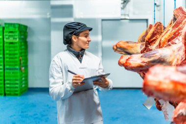 Üniformalı kadın ve panoya yazı yazan şapka et işleme fabrikasındaki malları kontrol ediyor.