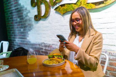 Beautiful latin woman wearing business casual clothes enjoying tasty burger and texting on the phone clipart