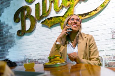 Stylish latin beauty blonde woman talking to the phone eating burger at restaurant clipart