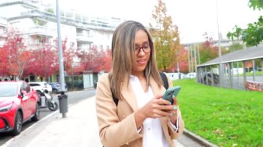 Latin kadın, günlük iş kıyafetleri içinde şehirde cep telefonuyla video görüşmesi yaparken kameraya el sallıyor.