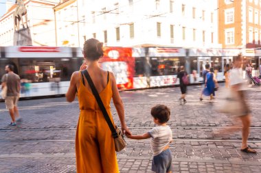 Yoğun bir caddede çocuğunu tutan bir kadın. Kadın sarı bir elbise giyiyor ve bir el çantası tutuyor. Çocuk beyaz bir gömlek ve mavi bir şort giyiyor. Etrafta dolaşan bir sürü insan var. freiburg im breisgau; freiburg