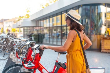 Sarı elbiseli bir kadın kırmızı bisiklete biniyor. O bir hasır şapka ve siyah bir çanta giyiyor.