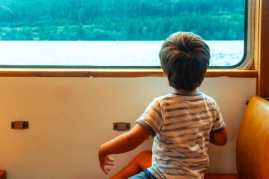 Trenin camında oturan genç bir çocuk denize bakıyor. Çocuk çizgili bir gömlek ve şort giyiyor.