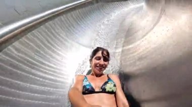 Bir kadın su kaydırağına uzanmış, gülümsüyor ve eğleniyor. Kaydırak gümüşten ve o da bir su parkının parçası.