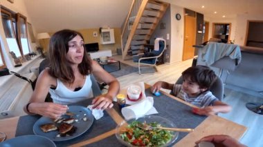 Bir kadın ve küçük bir çocuk yemek masasında birlikte yemek yiyorlar. Kadın salata yerken, oğlan bir sandviç yiyor. Masa tabaklar, kaseler ve çatal bıçaklarla dolu.