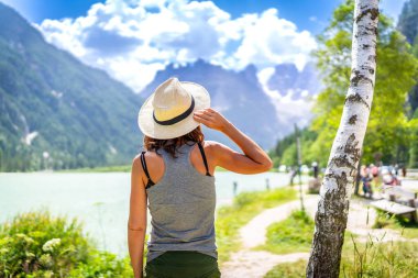 Woman wearing straw hat enjoying the view of the beautiful lago di landro with the dolomites in the background on a summer day, italy clipart