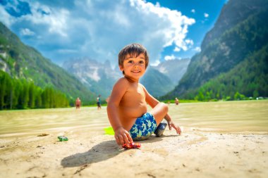 Smiling child playing with a toy car on the shore of lake landro, also called durrensee, in the italian dolomites, enjoying the summer holidays clipart