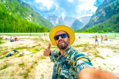 Tourist wearing sunglasses and straw hat gesturing thumbs up while taking a selfie at lago di landro with tourists enjoying summer in the dolomites, italy clipart