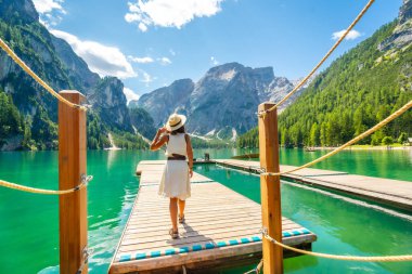 Braies Gölü 'ndeki ahşap iskelede yürüyen kadın, turkuaz suların ve güneşli bir yaz gününde dolomitlerin büyüleyici dağ manzarasının tadını çıkarıyor.