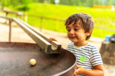 Meraklı bir çocuk oyun parkında yuvarlanan tahta bir topla eğleniyor, yerçekimini ve fiziği öğreniyor.