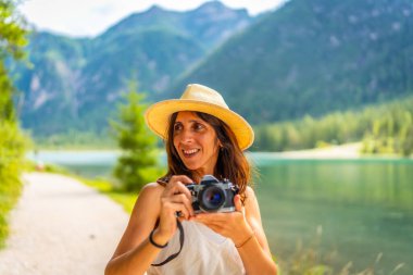 Hasır şapkalı gülümseyen turist yaz tatilinde Dolomitlerdeki Dobbiaco Gölü 'nde fotoğraf çekiyor.