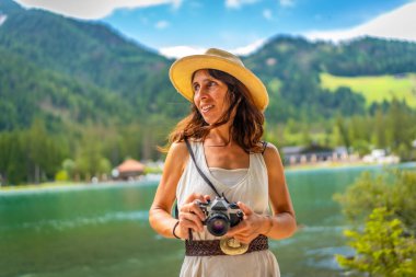 Woman wearing straw hat holding vintage camera enjoying breathtaking view of lake dobbiaco and surrounding mountains in the dolomites clipart